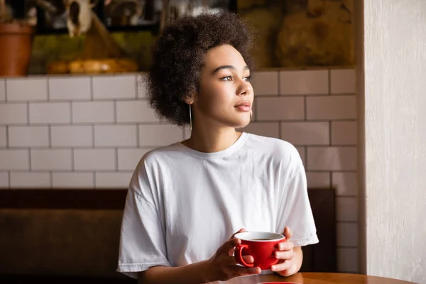 Thoughtful african american woman in white t-shirt holding cup — Stockfoto