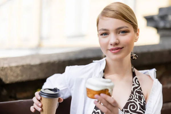 Junge blonde Frau mit Cupcake und Pappbecher lächelt draußen in die Kamera — Stockfoto