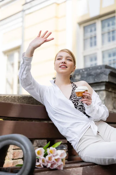 Smiling woman with cupcake waving hand while sitting on bench — Stock Photo