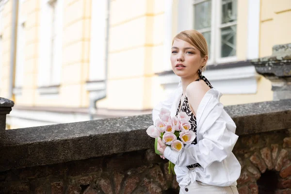 Stylish woman with tulips looking away near stone fence on street — стоковое фото