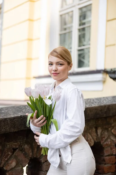 Mujer joven positiva sosteniendo tulipanes blancos y sonriendo al aire libre - foto de stock