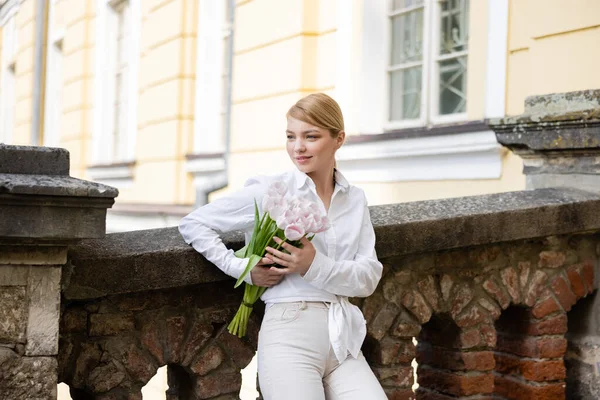 Young woman in stylish clothes holding white tulips while leaning on stone fence — стоковое фото