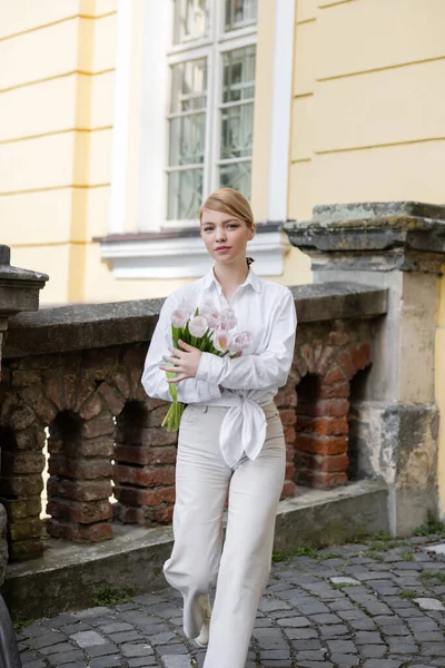 Mulher jovem e elegante com tulipas andando perto de cerca de pedra — Fotografia de Stock