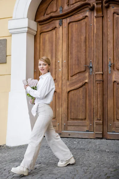 Piena vista lunghezza della donna elegante con tulipani a piedi vicino edificio con porte in legno — Foto stock