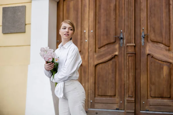 Hübsche junge Frau mit Tulpen, die in der Nähe verschwommener Holztüren wegschaut — Stockfoto