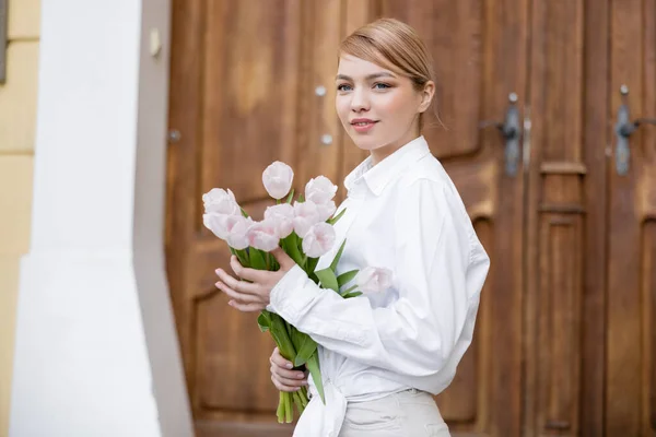 Femme heureuse en chemise blanche tenant bouquet de tulipes sur rue urbaine — Photo de stock