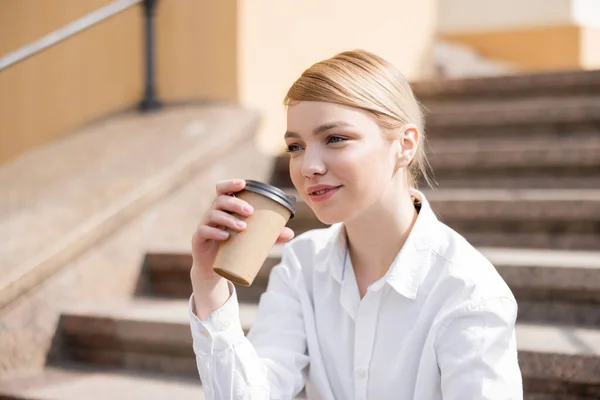 Pleased woman in white shirt holding coffee to go outdoors — Stock Photo