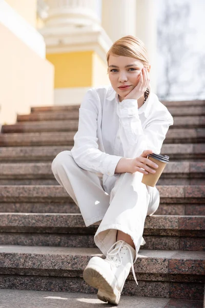 Stylish blonde woman with paper cup looking at camera while sitting on stairs — Stock Photo