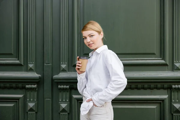 Jeune femme en chemise blanche debout avec la main dans la poche et le café pour aller près du mur gris — Photo de stock