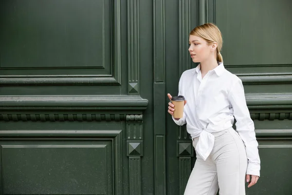 Bella donna in camicia bianca e pantaloni in piedi con tazza di carta vicino alla parete grigia — Foto stock