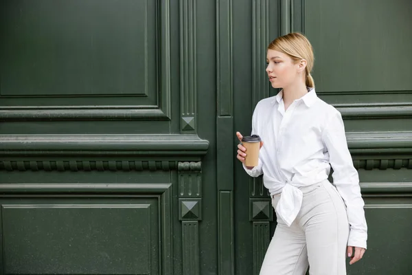 Bonita mujer en camisa blanca y pantalones de pie con taza de papel cerca de la pared gris - foto de stock