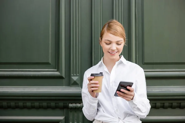 Joyeuse jeune femme avec boisson à emporter en regardant le téléphone mobile près du mur gris — Stock Photo