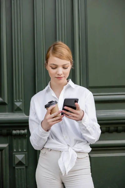 Mujer en camisa blanca sosteniendo la taza de papel y usando el teléfono celular cerca de la pared gris - foto de stock