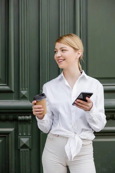 Femme heureuse regardant loin tout en se tenant debout avec smartphone et café pour aller près du mur gris — Photo de stock