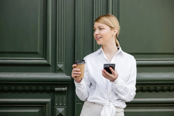 Heureuse jeune femme avec smartphone et café pour aller regarder loin près du mur gris — Photo de stock