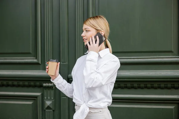 Mulher elegante com café para ir falar no smartphone perto da parede cinza — Fotografia de Stock