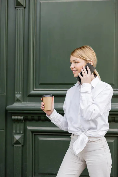 Mujer feliz llamando en el teléfono móvil, mientras que de pie con taza de papel cerca de la pared gris - foto de stock