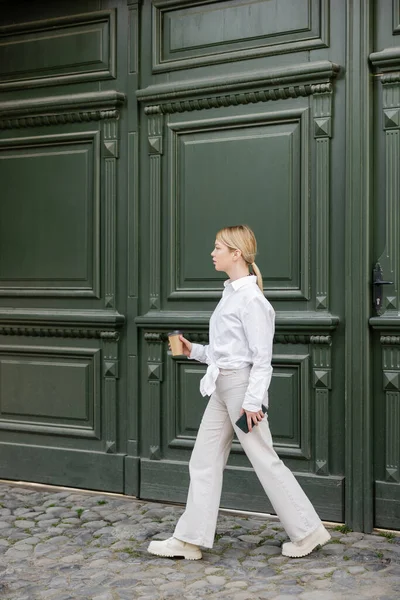 Vista completa de la mujer de moda con teléfono inteligente y café para ir caminando a lo largo de la pared gris - foto de stock