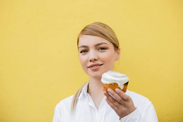 Happy young woman with cupcake looking at camera isolated on yellow — стоковое фото