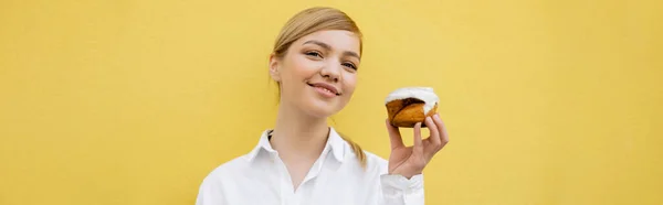 Joyful woman with cupcake smiling at camera isolated on yellow, banner — стоковое фото