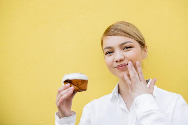 Femme heureuse tenant délicieux cupcake et essuyant lèvres isolées sur jaune — Photo de stock