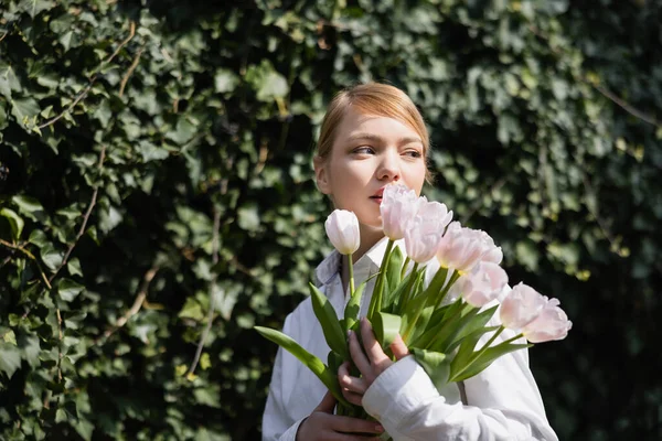 Jeune femme avec des tulipes blanches regardant loin tout en se tenant près de lierre flou — Photo de stock