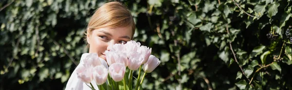 Femme souriante blonde visage obscurcissant avec des tulipes blanches près de lierre vert, bannière — Photo de stock