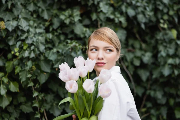 Giovane donna bionda con tulipani bianchi sorridenti vicino edera verde all'aperto — Foto stock