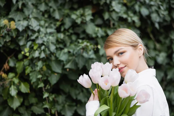 Charmante Frau mit weißen Tulpen lächelt in die Kamera in der Nähe von verschwommenem Efeu — Stockfoto