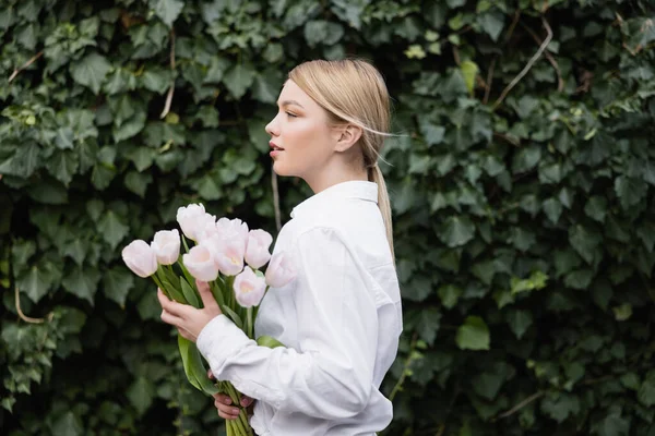 Side view of blonde woman with white tulips standing near green ivy — Stock Photo