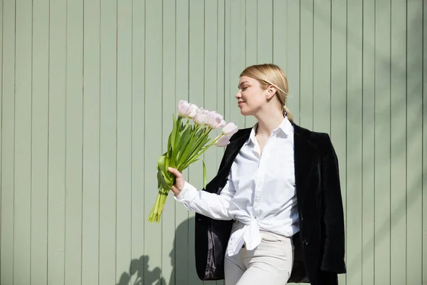 Mujer complacida en camisa blanca y chaqueta negra de pie con tulipanes cerca de la pared gris — Stock Photo