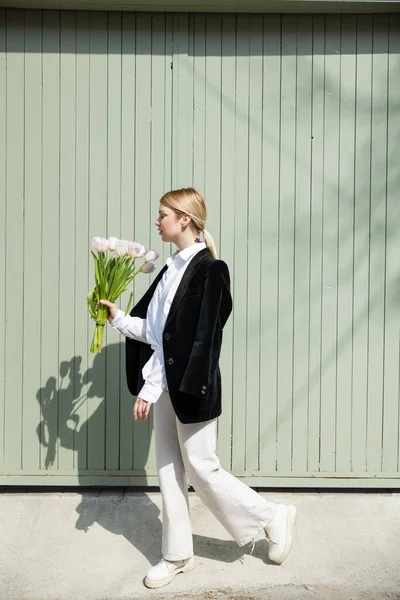 Side view of stylish woman with white tulips walking along grey wall — Stock Photo
