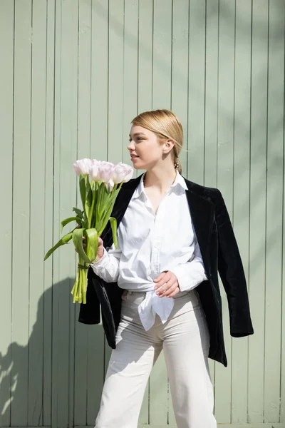 Mulher loira de pé perto da parede cinza com buquê de tulipas brancas — Fotografia de Stock