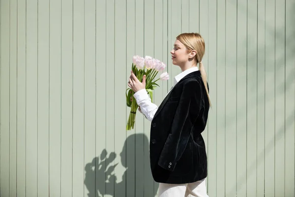 Vista lateral de mujer joven en chaqueta negra sosteniendo tulipanes blancos cerca de la pared gris - foto de stock