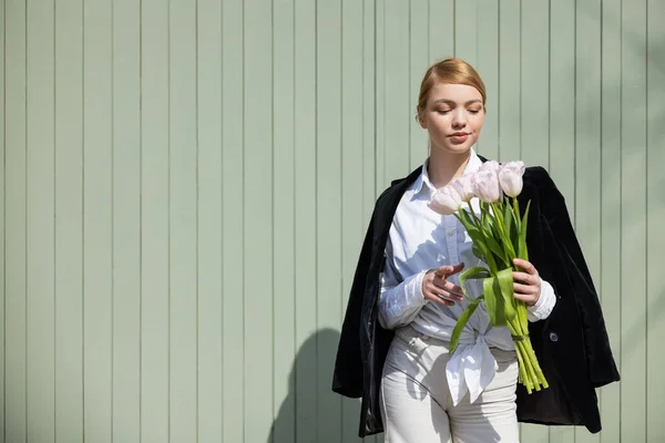 Mulher bonita em blazer preto de pé perto da parede cinza com tulipas brancas — Fotografia de Stock