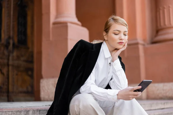 Jeune femme en chemise blanche et blazer noir en utilisant smartphone sur la rue urbaine — Photo de stock