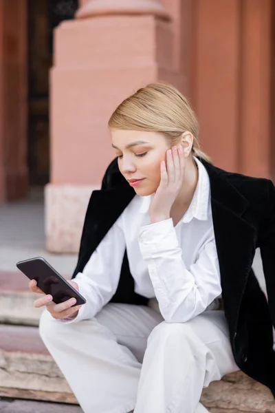 Femme blonde élégante utilisant un téléphone portable tout en étant assis à l'extérieur — Photo de stock