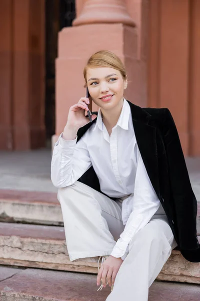Pleased young woman sitting on street and talking on mobile phone — стоковое фото