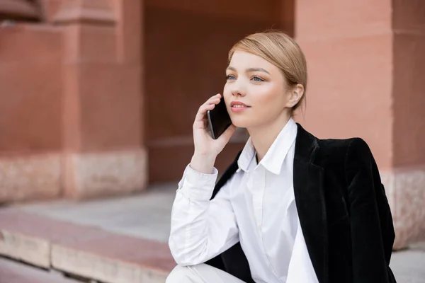 Pretty young woman calling on smartphone while sitting on city street — Stock Photo