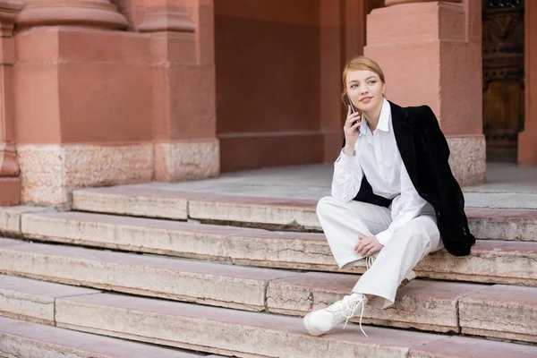 Full length view of stylish woman sitting on stairs and talking on smartphone — стоковое фото