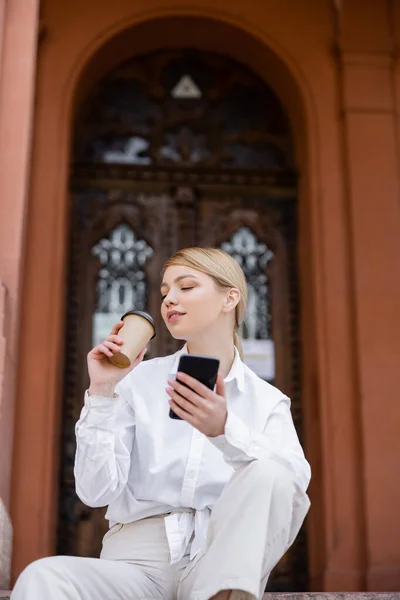 Blonde woman with smartphone drinking coffee near blurred building — Stock Photo