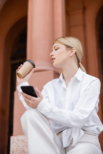 Femme blonde en chemise blanche assise avec du café à emporter et smartphone à l'extérieur — Photo de stock