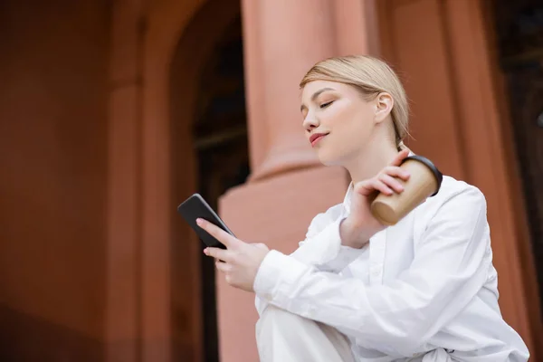 Jovem mulher em camisa branca usando smartphone enquanto segurando copo de papel ao ar livre — Fotografia de Stock