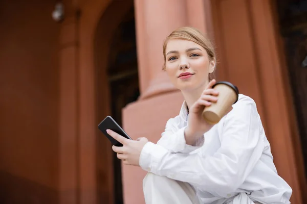 Mujer rubia con taza de papel y teléfono móvil mirando a la cámara al aire libre - foto de stock