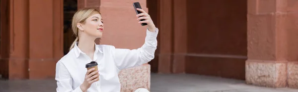 Charming woman with takeaway drink taking selfie on smartphone outdoors, banner — Stock Photo