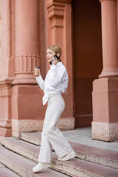 Jeune femme à la mode avec du café pour aller marcher dans les escaliers et parler sur téléphone portable — Photo de stock