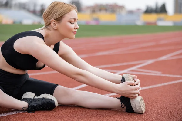 Young sportswoman stretching leg while sitting on stadium — Stock Photo