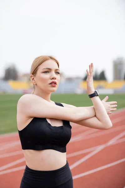 Mujer joven en sujetador deportivo negro calentándose al aire libre y mirando hacia otro lado - foto de stock