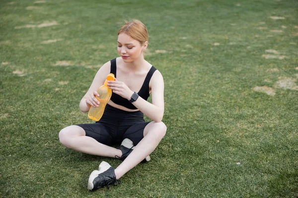 Young sportswoman sitting on stadium and opening sports bottle — Stock Photo