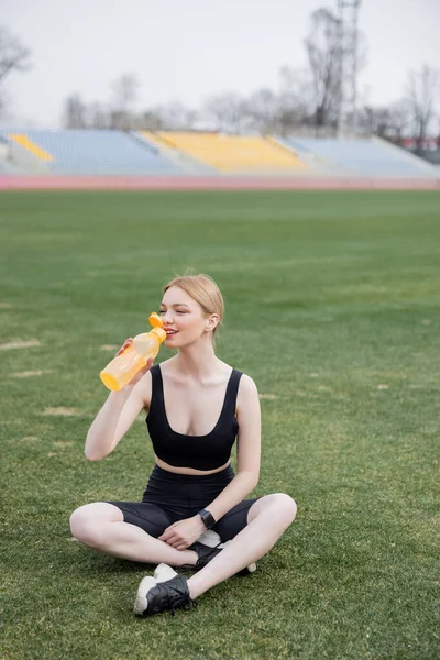 Donna sorridente seduta sul campo sportivo e che beve dalla bottiglia sportiva — Foto stock